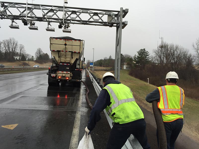 Workers placing material along a highway guardrail beside a truck in rainy weather.
